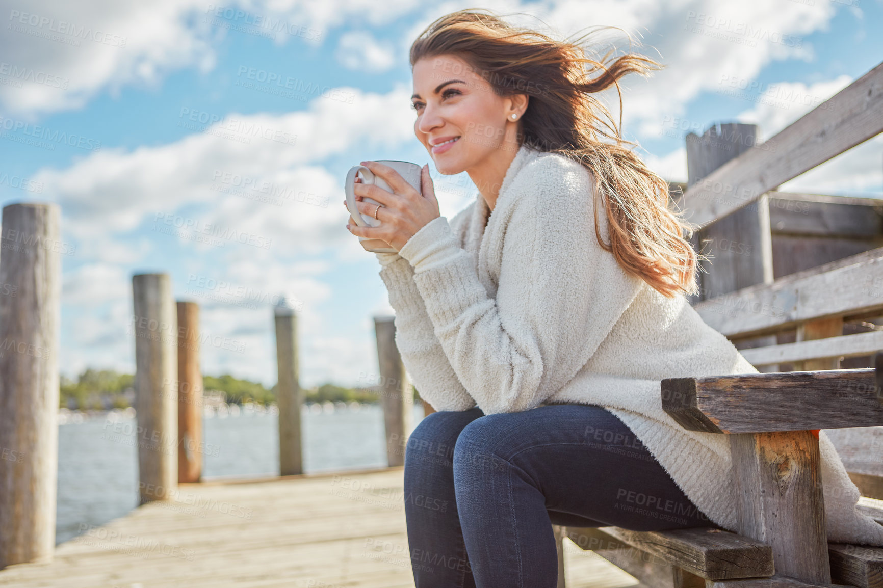 Buy stock photo Shot of a beautiful young woman enjoying a warm beverage while relaxing on a bench at a lake