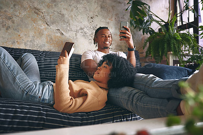 Buy stock photo Shot of a young woman reading a book while her boyfriend uses a cellphone at home