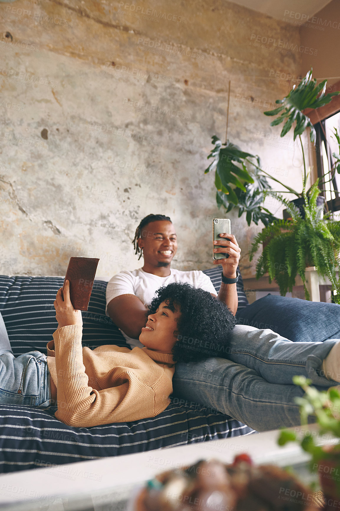 Buy stock photo Shot of a young woman reading a book while her boyfriend uses a cellphone at home