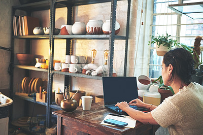 Buy stock photo Cropped shot of an unrecognizable business owner sitting alone in her pottery studio and using her laptop