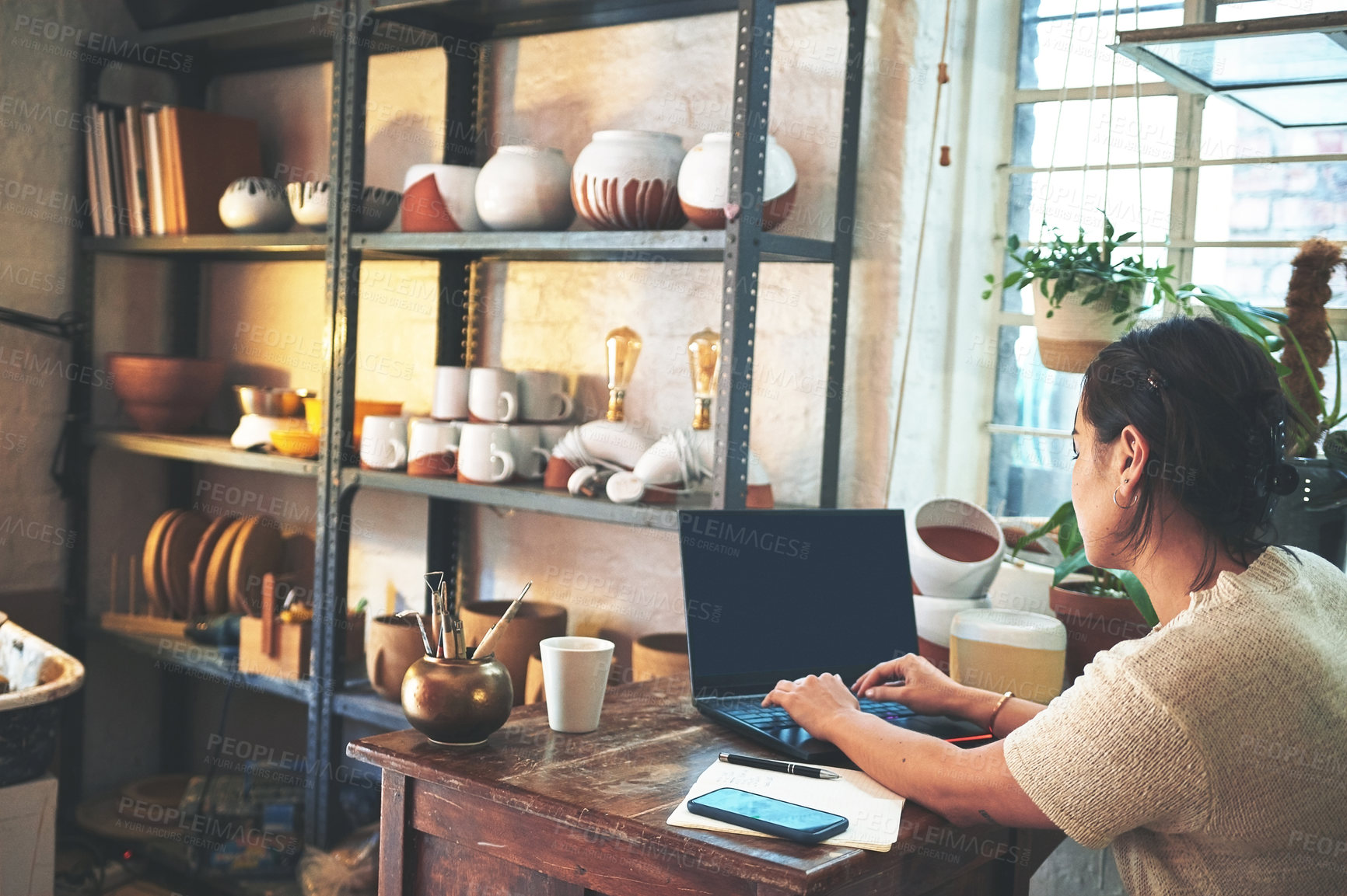 Buy stock photo Cropped shot of an unrecognizable business owner sitting alone in her pottery studio and using her laptop