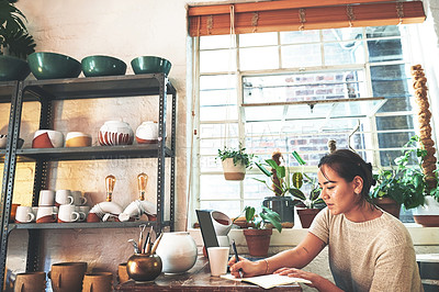 Buy stock photo Cropped shot of an attractive young business owner sitting alone in her pottery studio and making notes