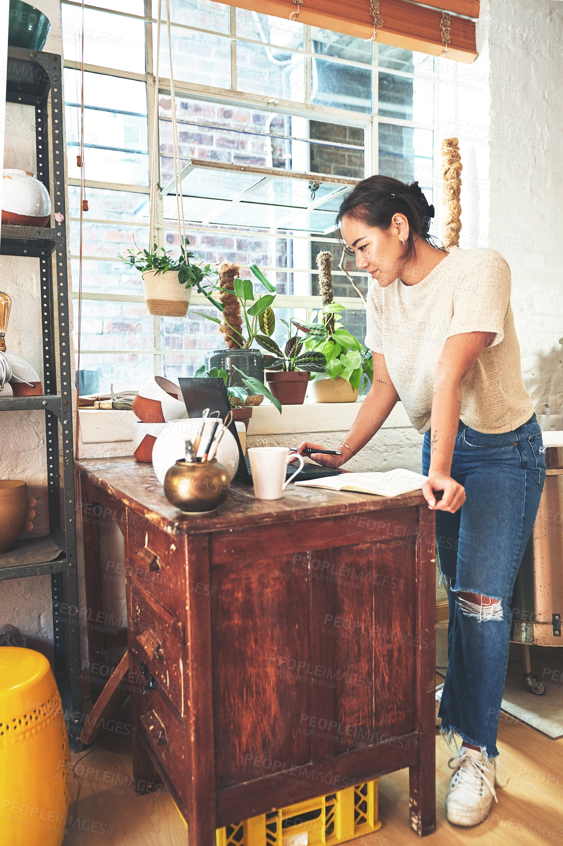 Buy stock photo Full length shot of an attractive young business owner standing alone in her pottery studio and using her laptop