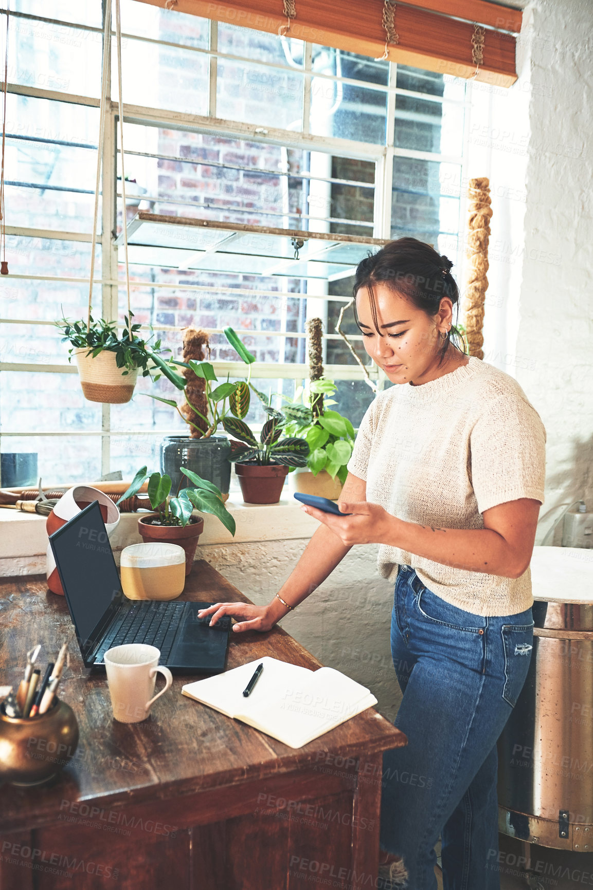 Buy stock photo Cropped shot of an attractive young business owner standing and using her laptop and cellphone in her pottery studio