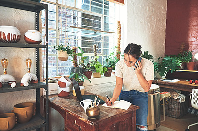 Buy stock photo Cropped shot of an attractive young business owner standing and using her cellphone while taking notes in her pottery studio