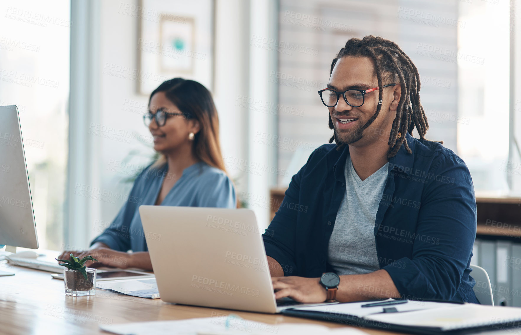 Buy stock photo Shot of a young businessman working on a laptop in an office with his colleague in the background