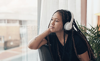 Buy stock photo Shot of a young woman using headphones and looking thoughtfully out of a window at home