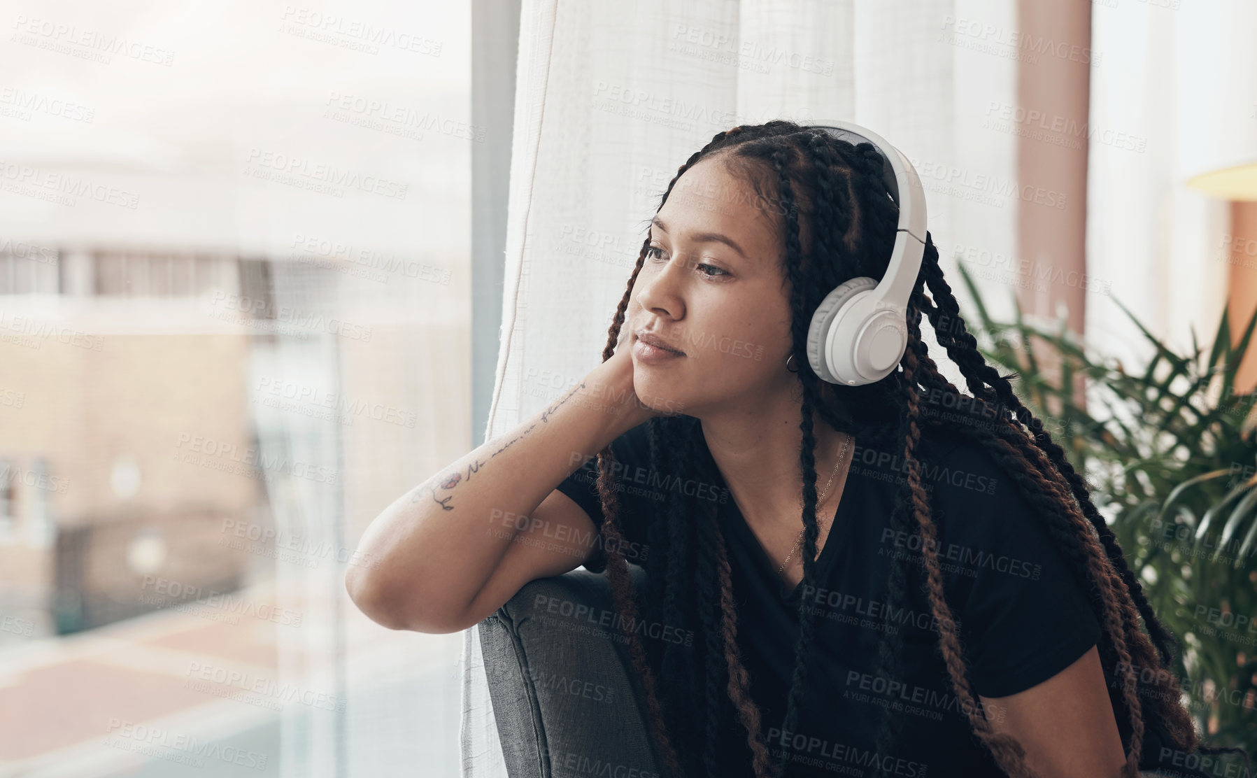 Buy stock photo Shot of a young woman using headphones and looking thoughtfully out of a window at home