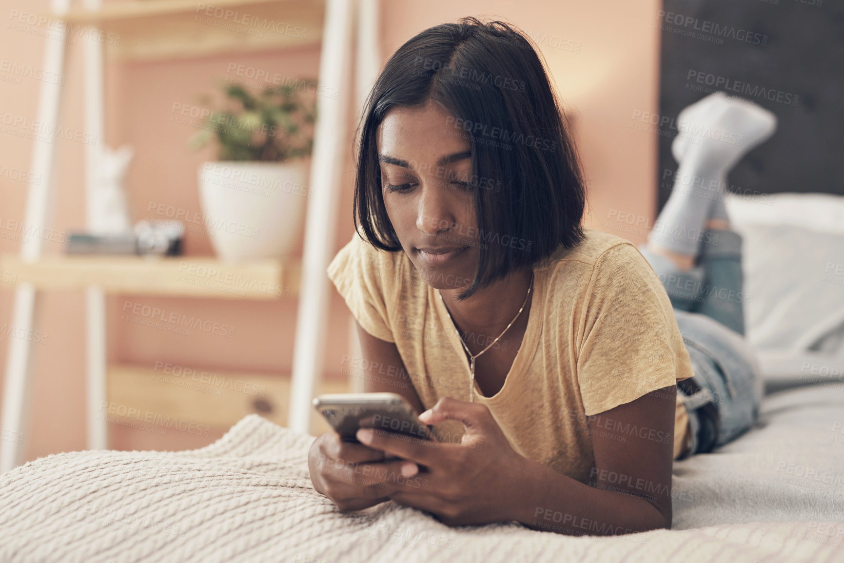 Buy stock photo Shot of a young woman using a smartphone while relaxing on her bed at home