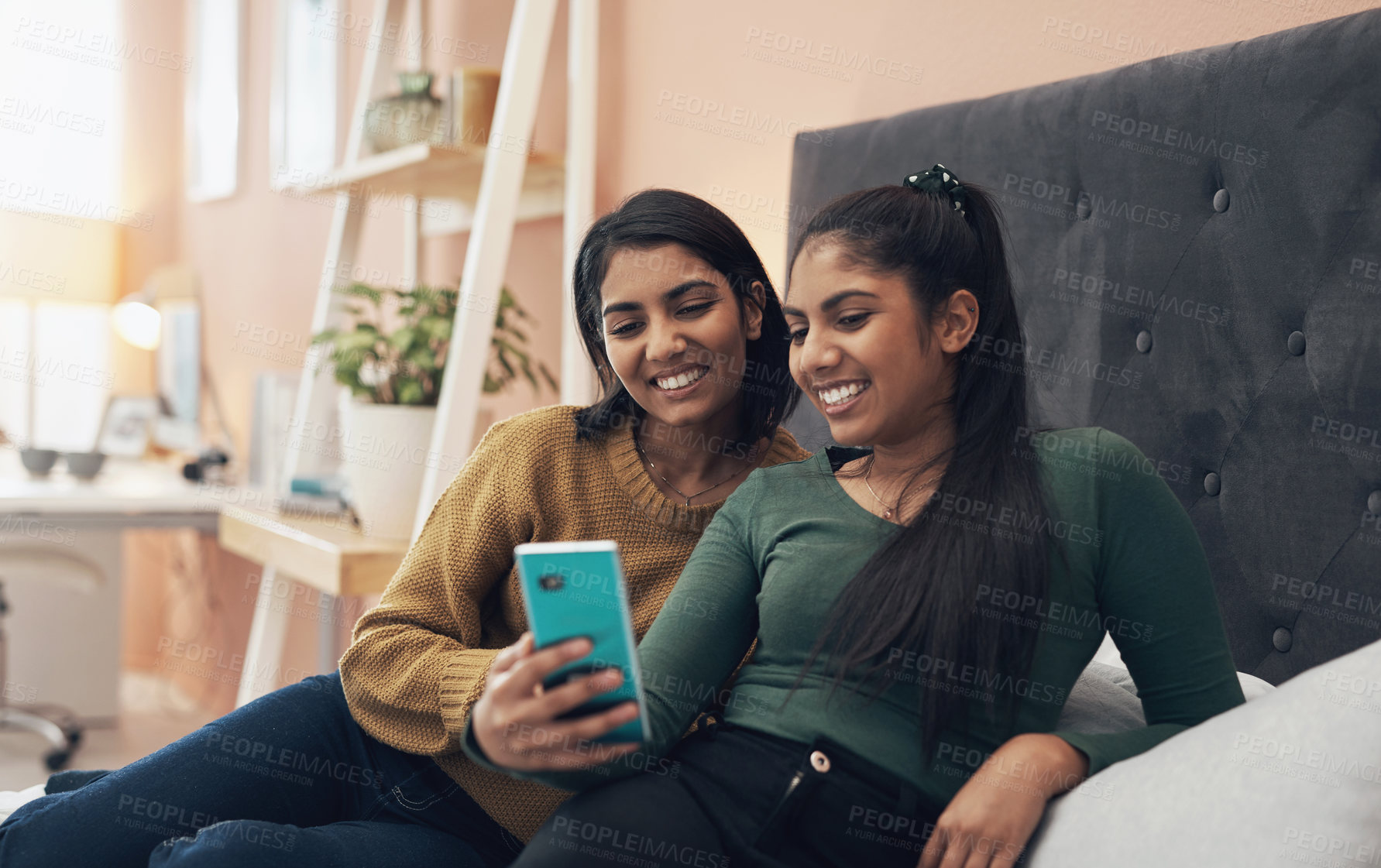 Buy stock photo Shot of two young women looking at something on a cellphone while sitting on a bed together