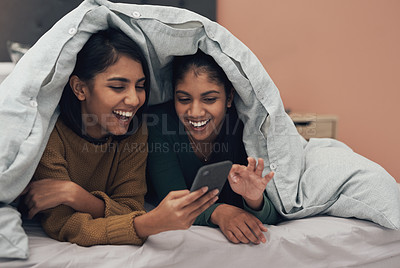 Buy stock photo Shot of two young women looking at something on a cellphone while lying on a bed