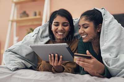Buy stock photo Shot of two young women using a digital tablet and a credit card while lying on a bed together