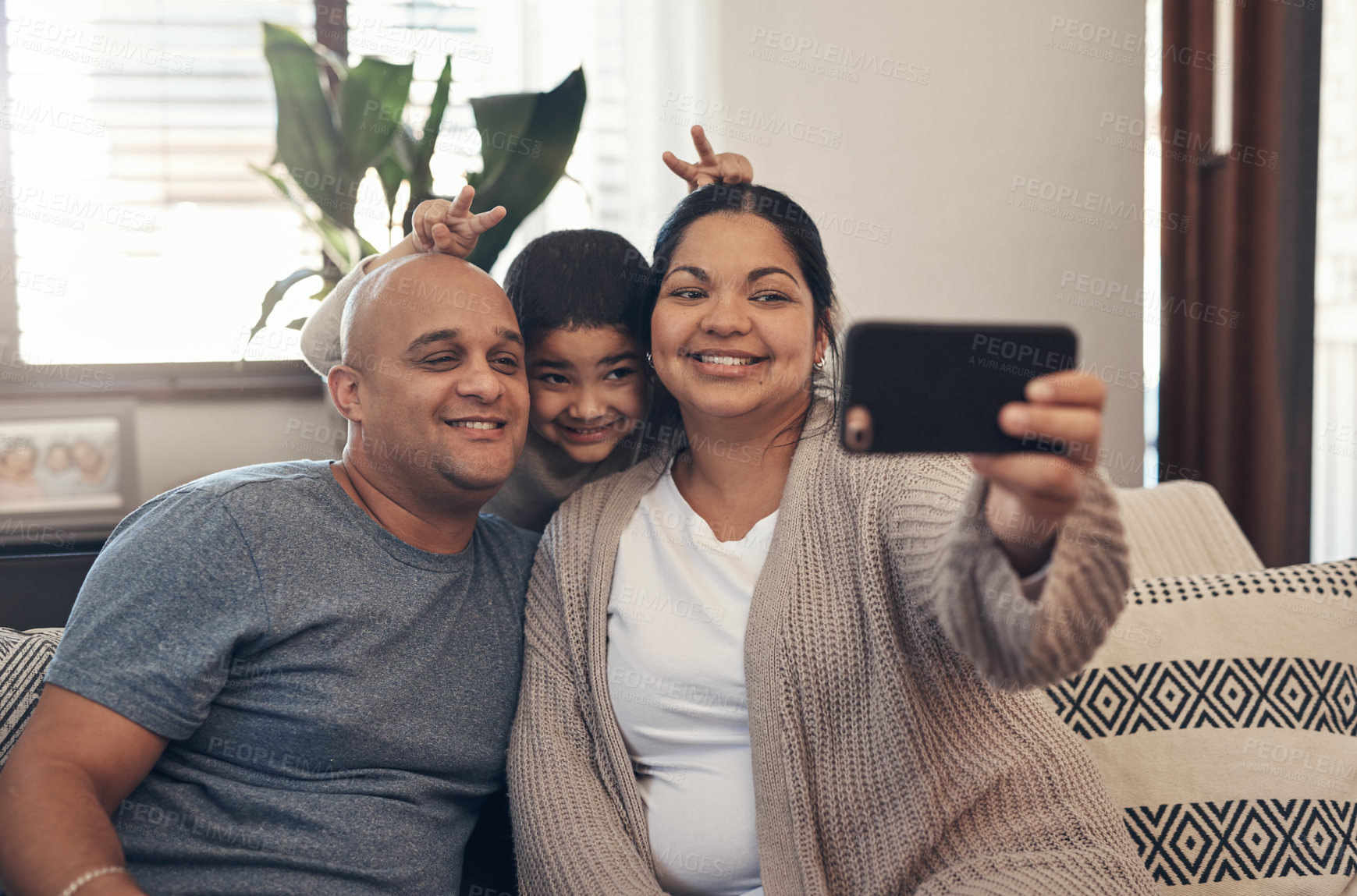 Buy stock photo Shot of a happy young family taking selfies with a smartphone on the sofa at home