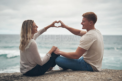 Buy stock photo Cropped shot of a couple forming a heart shape with their hands while sitting on the beach