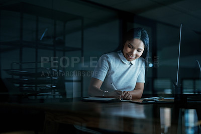 Buy stock photo Shot of a young businesswoman writing in a notebook and using a computer during a late night at work