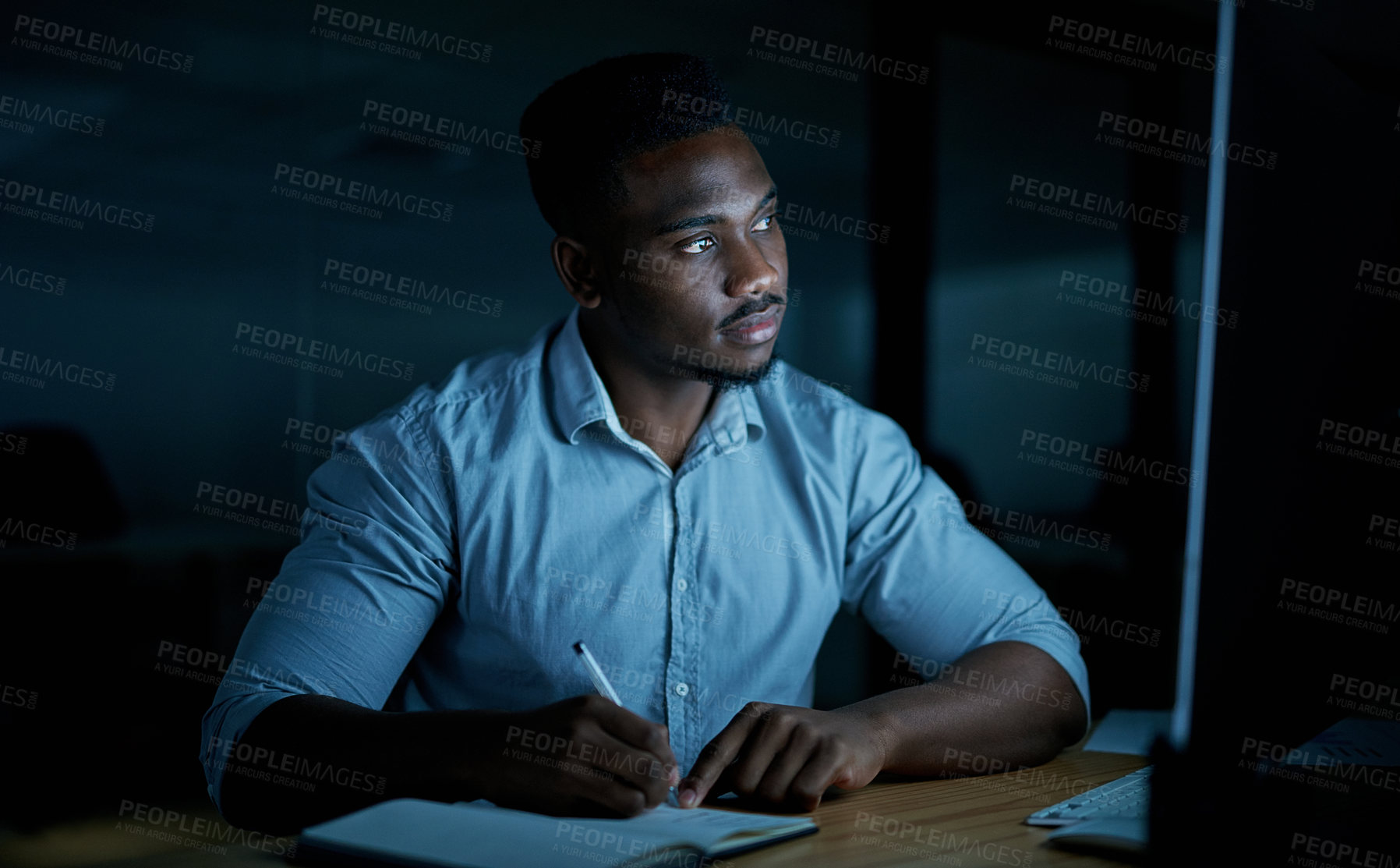 Buy stock photo Shot of a young businessman writing in a notebook and using a computer during a late night at work