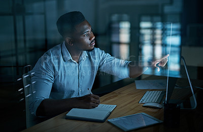 Buy stock photo Shot of a young businessman writing in a notebook and using a computer during a late night at work