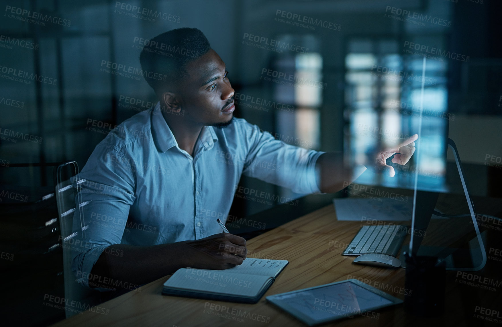 Buy stock photo Shot of a young businessman writing in a notebook and using a computer during a late night at work