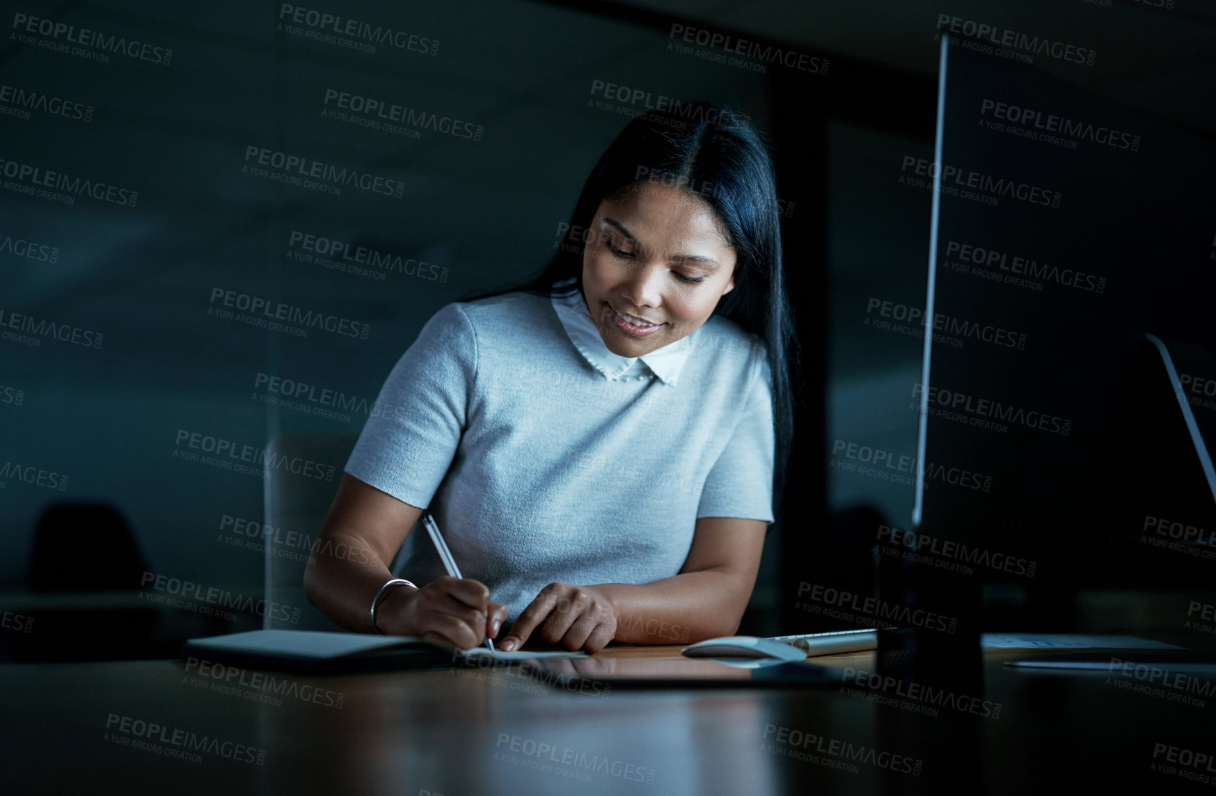 Buy stock photo Shot of a young businesswoman writing in a notebook and using a computer during a late night at work