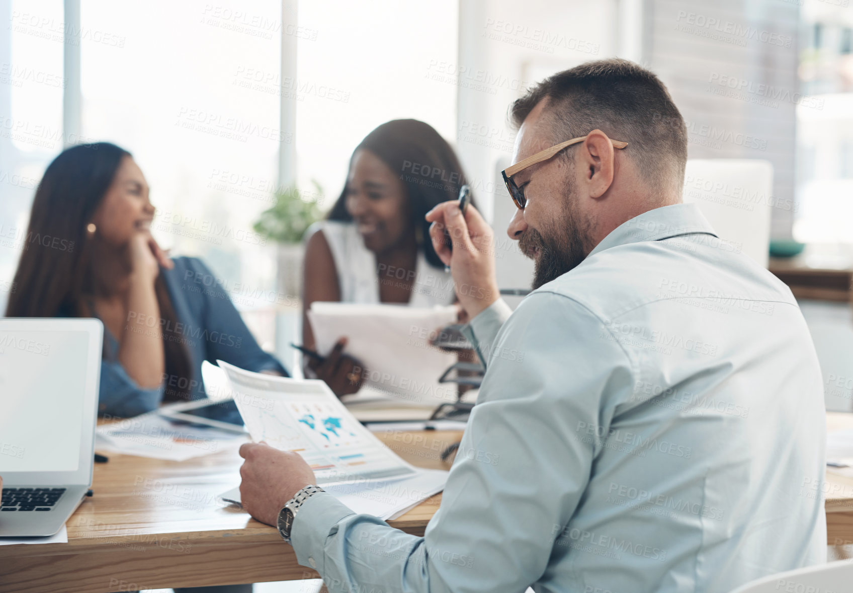 Buy stock photo Cropped shot of a handsome young businessman sitting and reading paperwork while his colleagues sit in the background