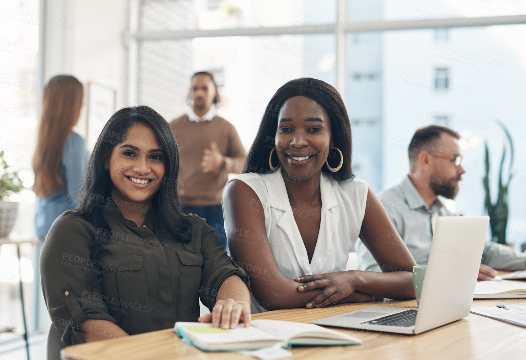 Buy stock photo Cropped portrait of two young businesswomen sitting together and using a laptop while their colleagues work in the background