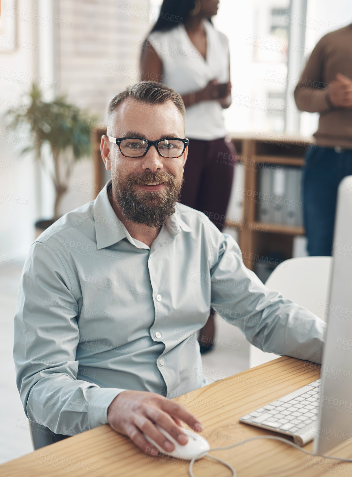 Buy stock photo Cropped portrait of a handsome young businessman sitting and using a computer while his colleagues work in the background