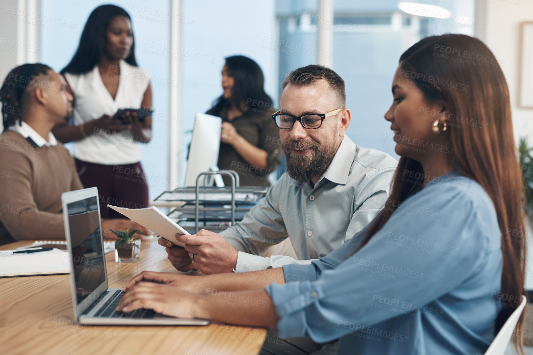 Buy stock photo Cropped shot of two young businesspeople sitting together and using a laptop while their colleagues work in the background