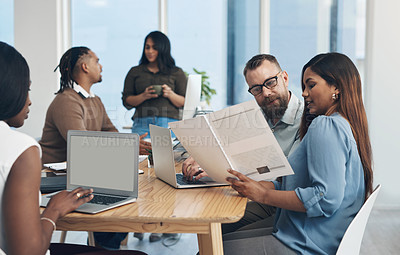 Buy stock photo Cropped shot of two young businesspeople sitting together and reading through paperwork while their colleagues work in the background