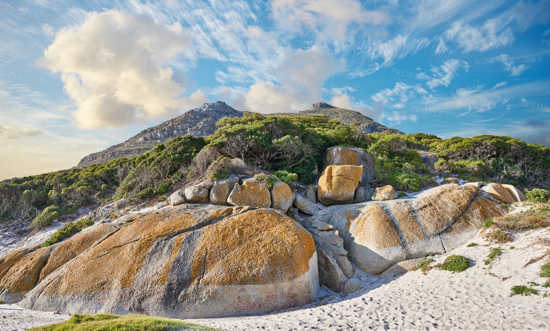 Buy stock photo Rocks and boulders on a coastal shore against a mountain background with lush green plants and shrubs below a cloudy blue sky. Beautiful view and tourist attraction in Cape Town, South Africa