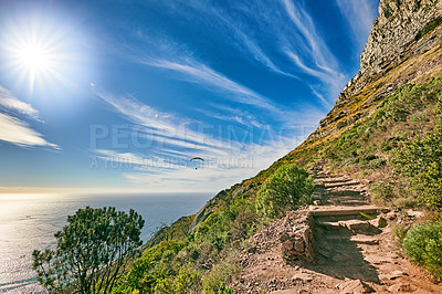 Buy stock photo A mountain trail with blue sky and lensflare over ocean. Landscape of mysterious dirt road for hiking on adventure walks along a beautiful scenic trail with lush shrubs in Cape Town, South Africa