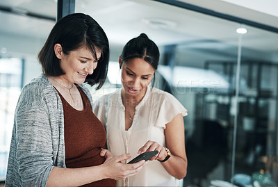 Buy stock photo Cropped shot of two young businesswoman standing together in the office and using a cellphone