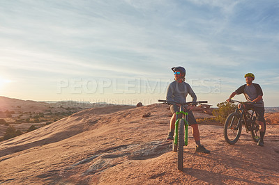 Buy stock photo Full length shot of two young male athletes mountain biking in the wilderness