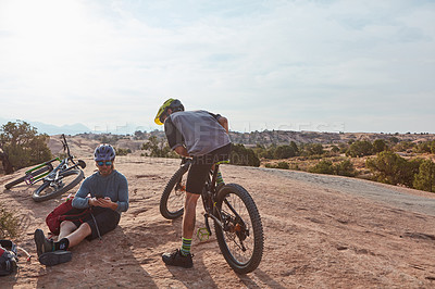 Buy stock photo Full length shot of two athletic men taking a break while mountain biking through the wilderness