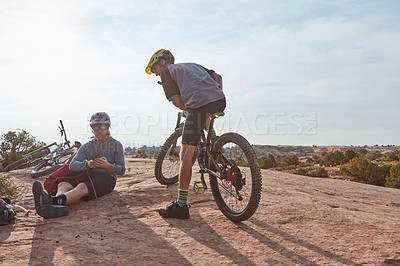 Buy stock photo Full length shot of two athletic men taking a break while mountain biking through the wilderness