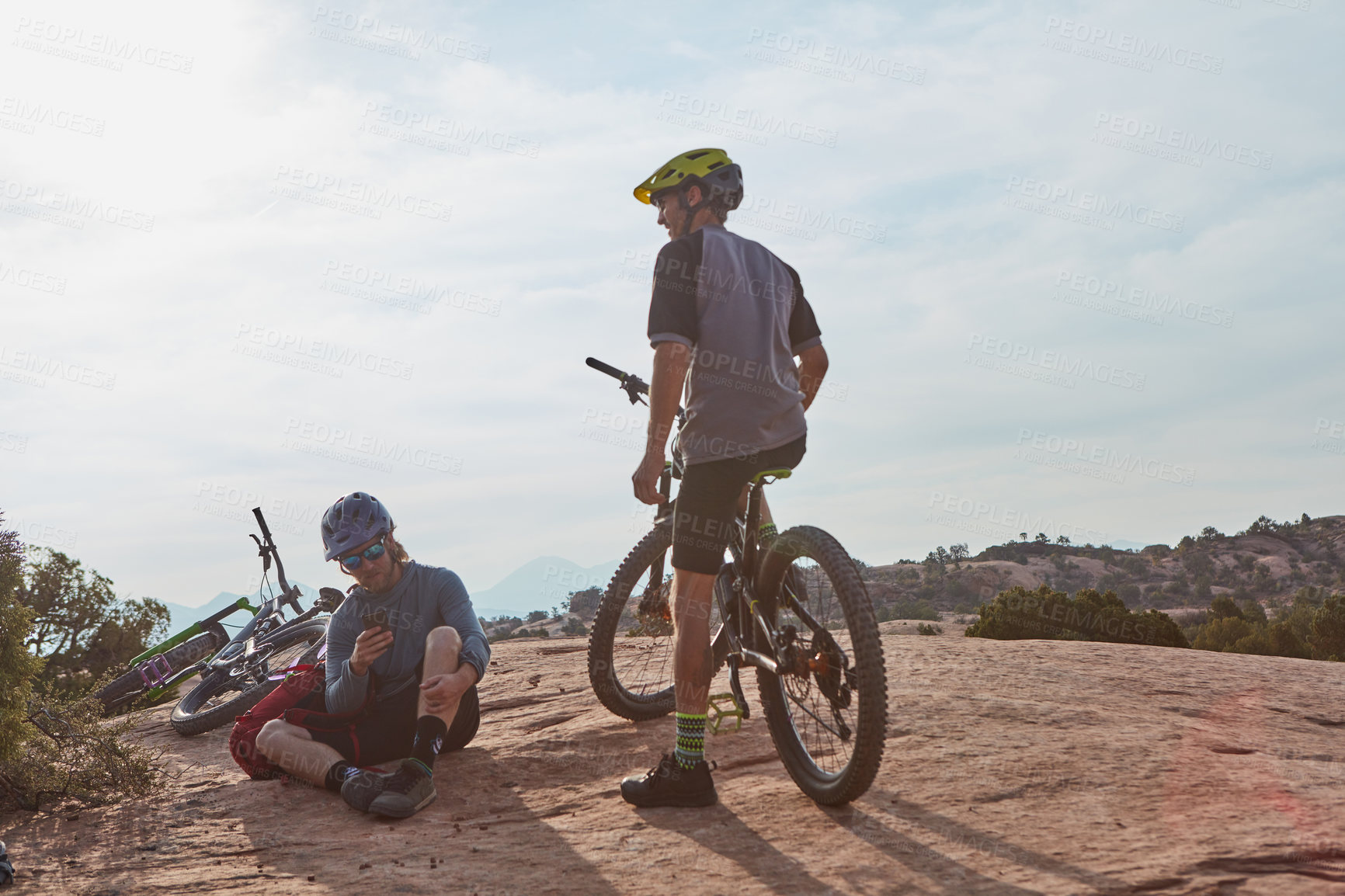 Buy stock photo Full length shot of two athletic men taking a break while mountain biking through the wilderness