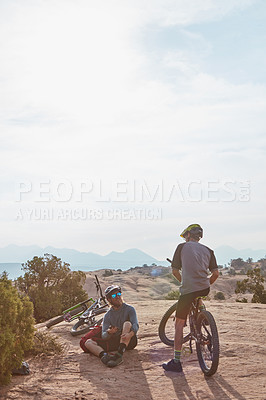 Buy stock photo Full length shot of two athletic men taking a break while mountain biking through the wilderness