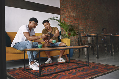 Buy stock photo Shot of two young men discussing something on a digital tablet while sitting together in a coffee shop