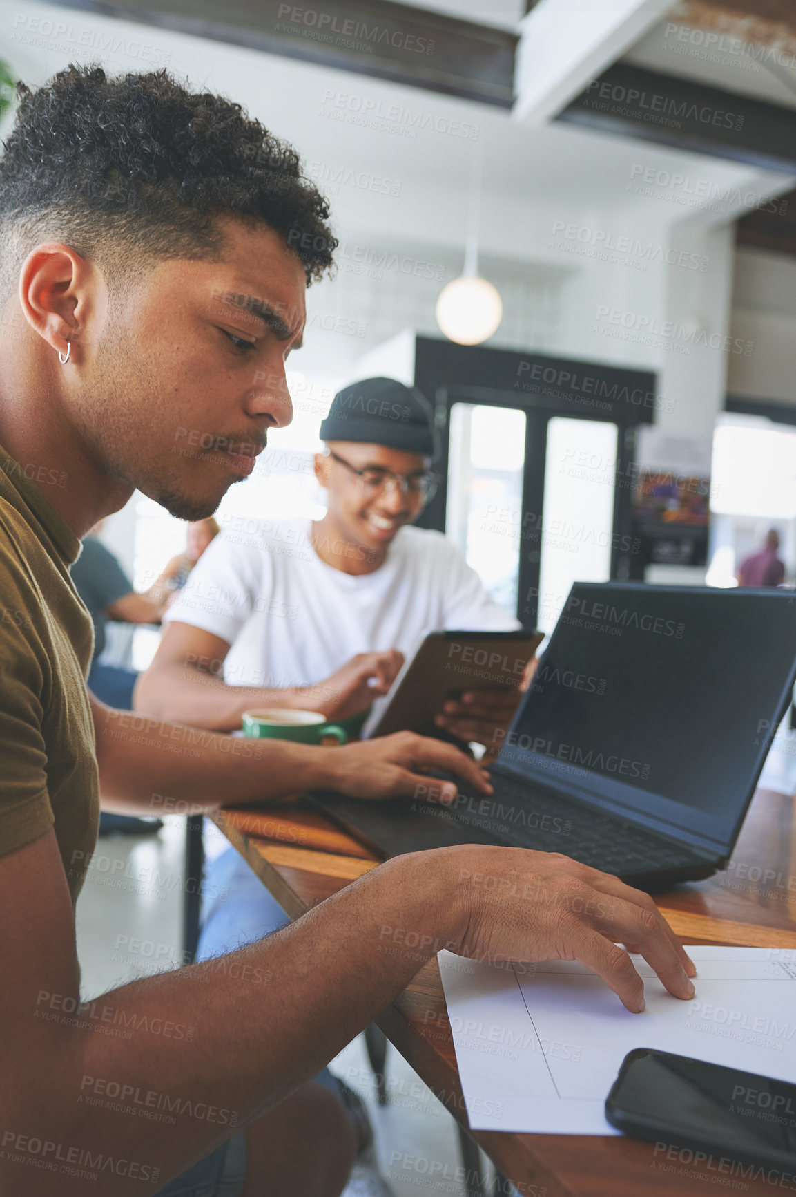 Buy stock photo Cropped shot of a handsome young man sitting with a friend and using a laptop while reading paperwork