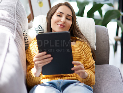 Buy stock photo Shot of a young woman using a digital tablet while relaxing on a sofa at home