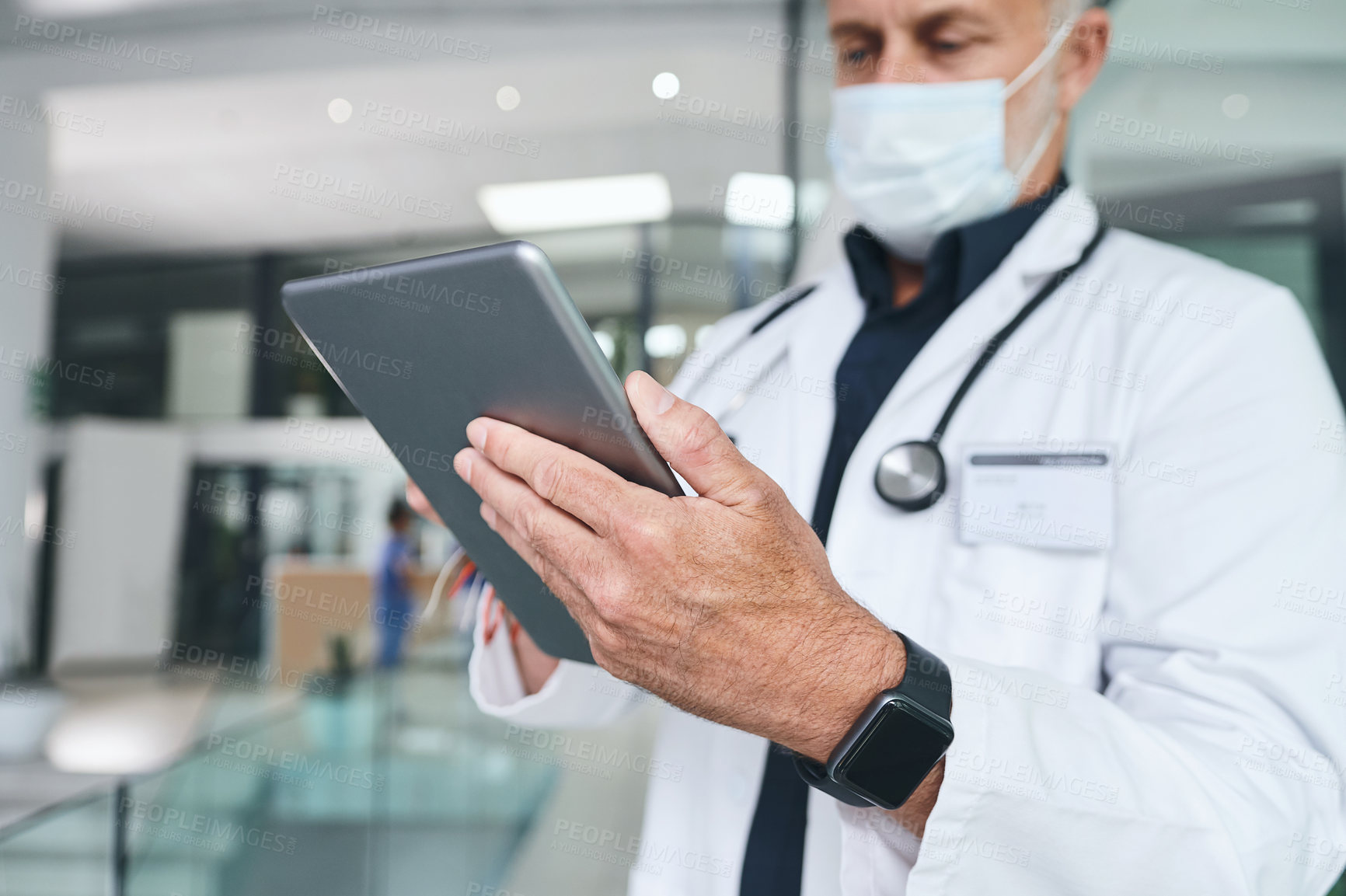 Buy stock photo Cropped shot of a handsome mature doctor standing and wearing a face mask while using a digital tablet