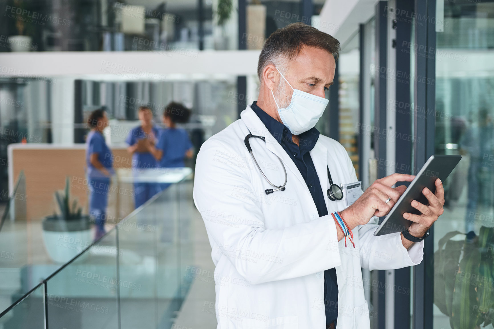 Buy stock photo Cropped shot of a handsome mature doctor standing and wearing a face mask while using a digital tablet