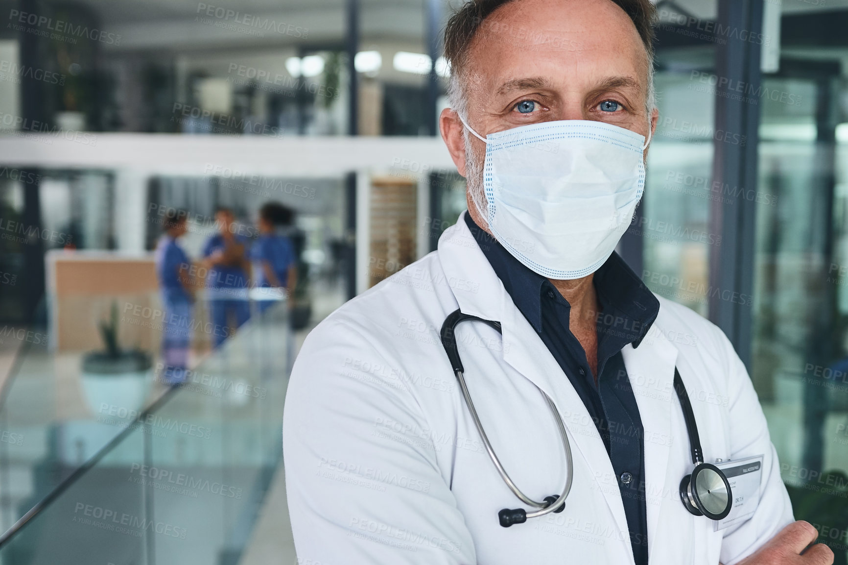 Buy stock photo Cropped portrait of a handsome mature doctor wearing a face mask and standing alone in the clinic