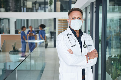 Buy stock photo Cropped portrait of a handsome mature doctor wearing a face mask and standing alone in the clinic