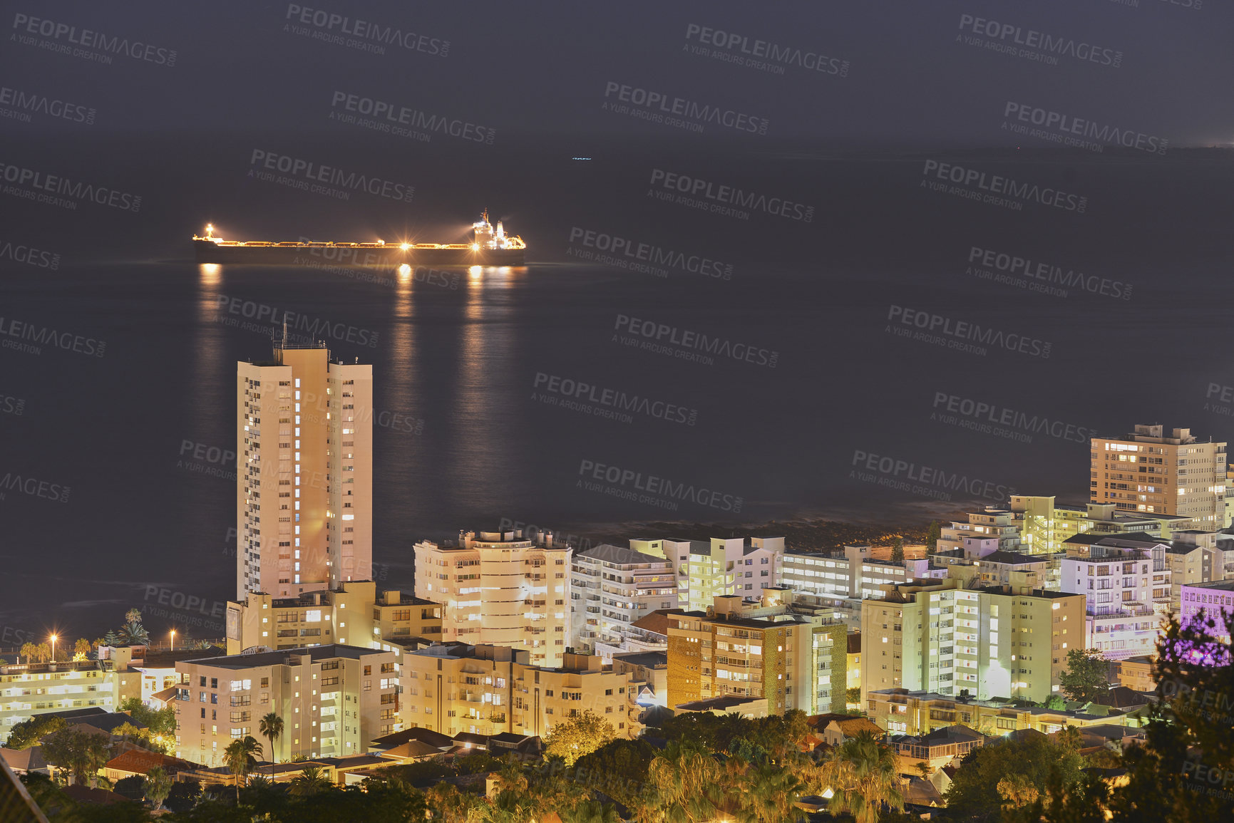 Buy stock photo View of skyscraper and city buildings lighting up the dark sky at night, alongside the ocean horizon. Beautiful scenic urban landscape in the evening with a seascape background with a ship at sea.