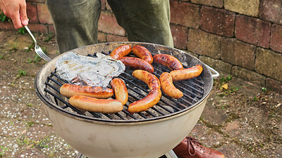 Buy stock photo Unrecognizable shot of a man barbecuing chicken and sausage outside in the rain