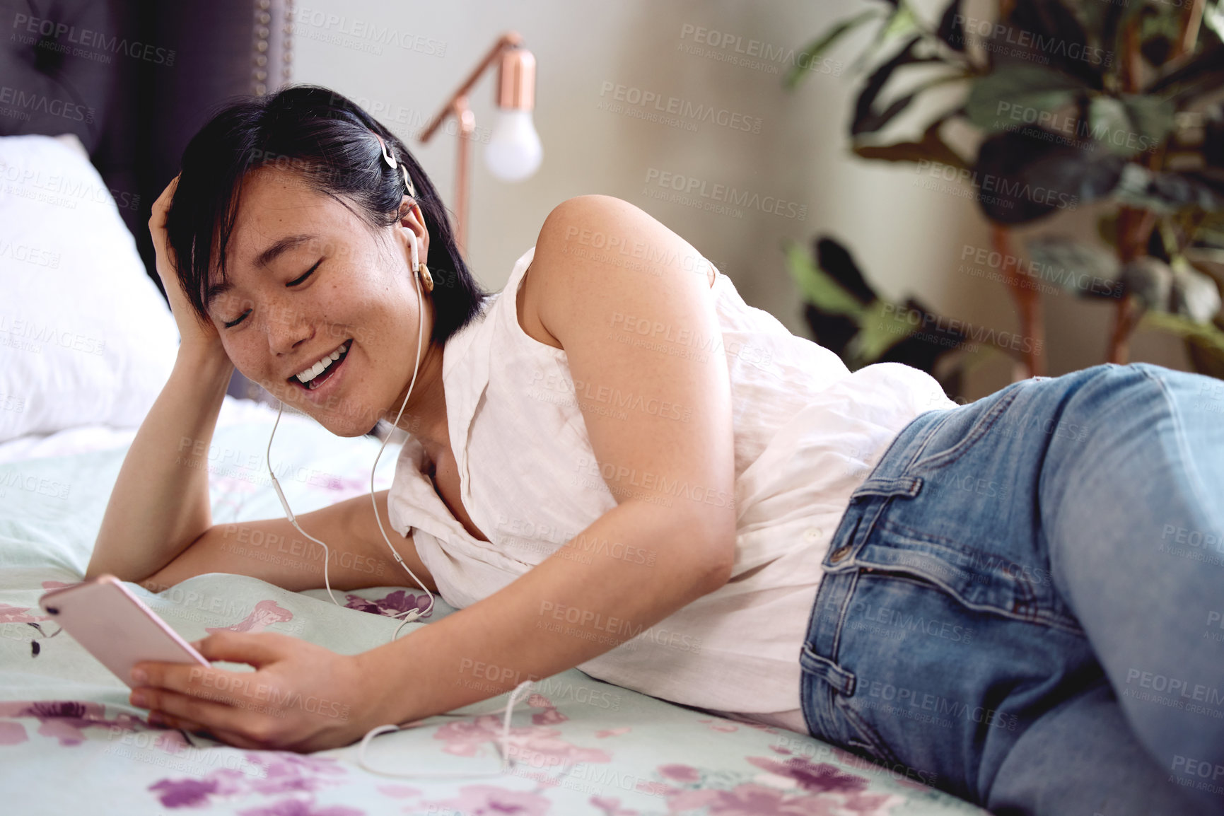 Buy stock photo Shot of a young woman lying on her bed listening to music on her smartphone at home