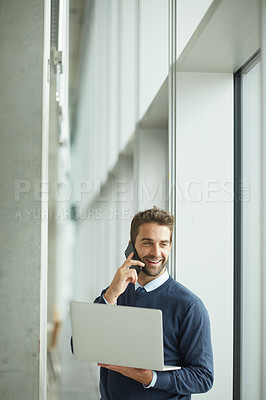 Buy stock photo Cropped shot of a handsome young businessman standing alone indoors and using his cellphone and laptop