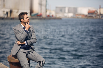 Buy stock photo Cropped shot of a handsome young businessman sitting alone by the harbour and looking contemplative during the day