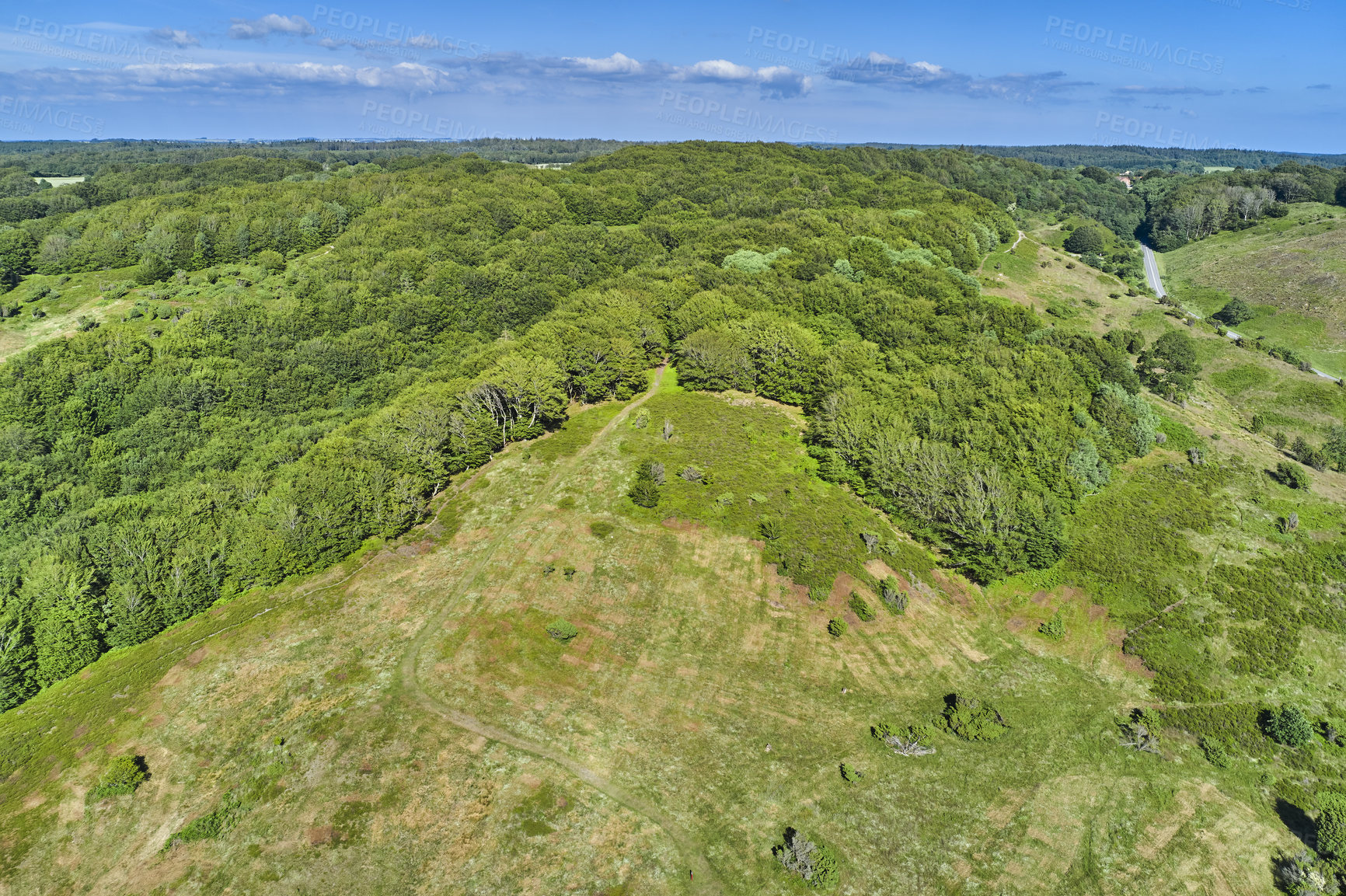 Buy stock photo Aerial view of a field at the top of a hill in the countryside, surrounded by green forestry and trees on a sunny spring or summer day. Scenery of nature with the blue sky on the horizon.