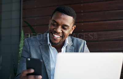 Buy stock photo Shot of a handsome young businessman sitting alone in the office while using his cellphone and laptop